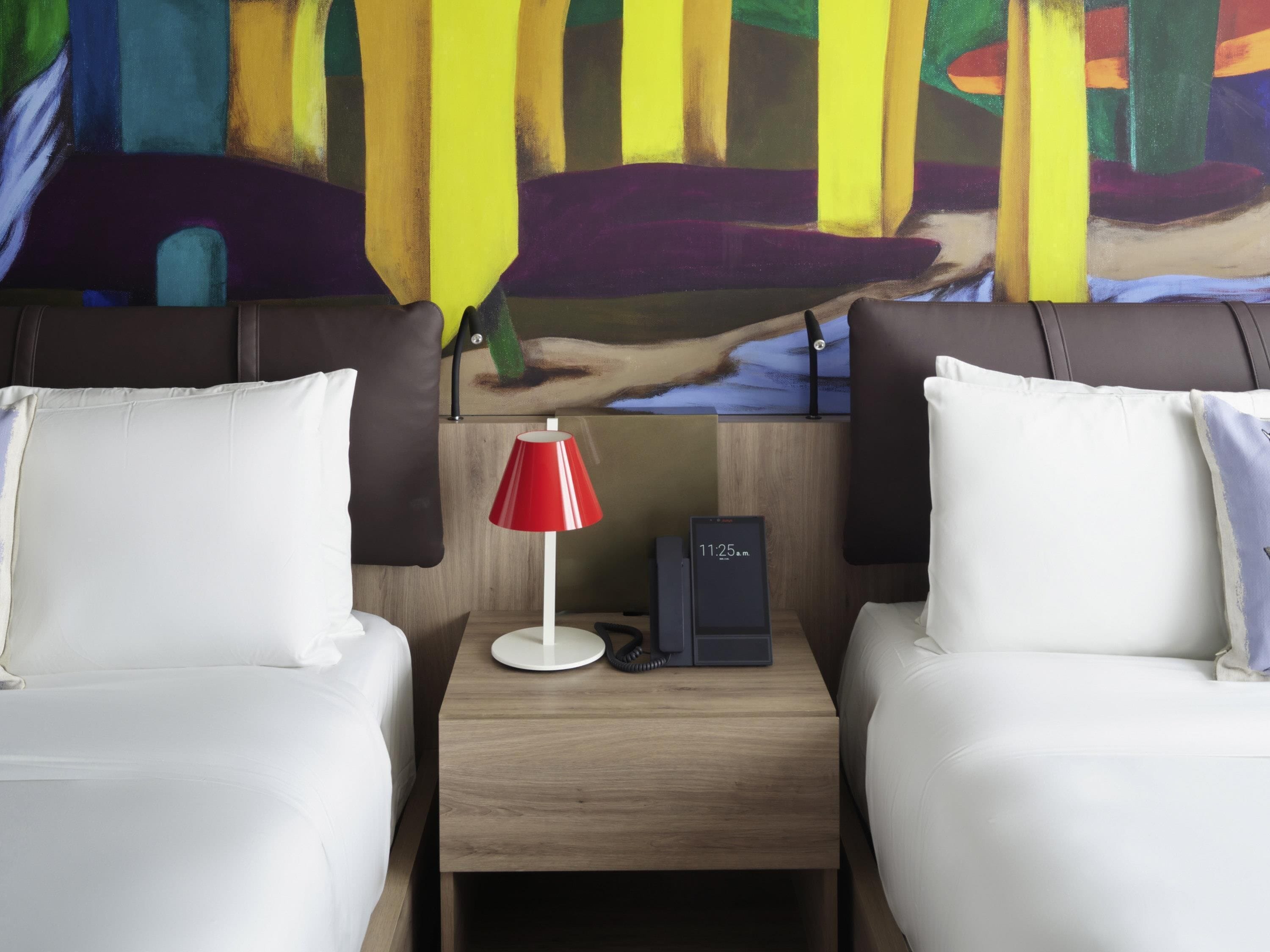 Een hotelkamer met twee bedden, een nachtkastje met een rode lamp, een klok en een schilderij aan de muur.