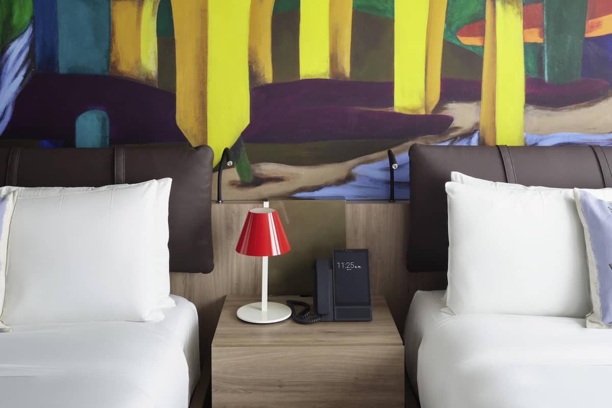 Een hotelkamer met twee bedden, een nachtkastje met een rode lamp, een klok en een schilderij aan de muur.