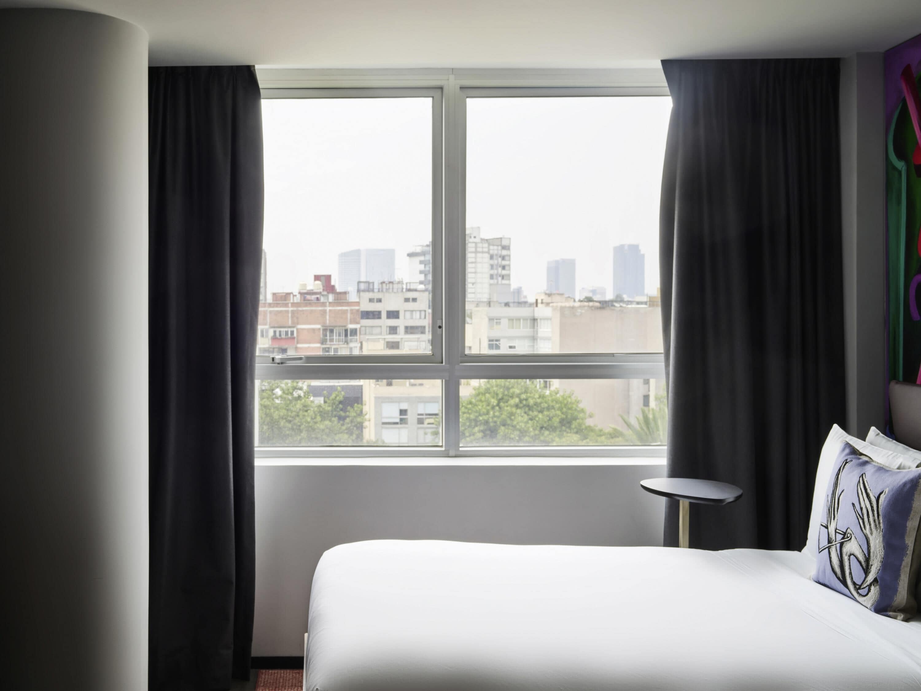 Habitación de hotel con una ventana grande, una cama, una mesita y vistas a la ciudad.
