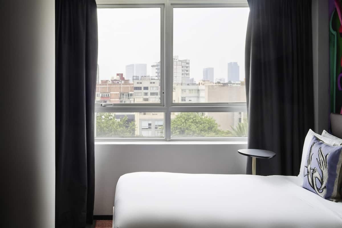 Hotellihuone, jossa on suuri ikkuna, sänky, pieni pöytä ja näkymä kaupunkiin.