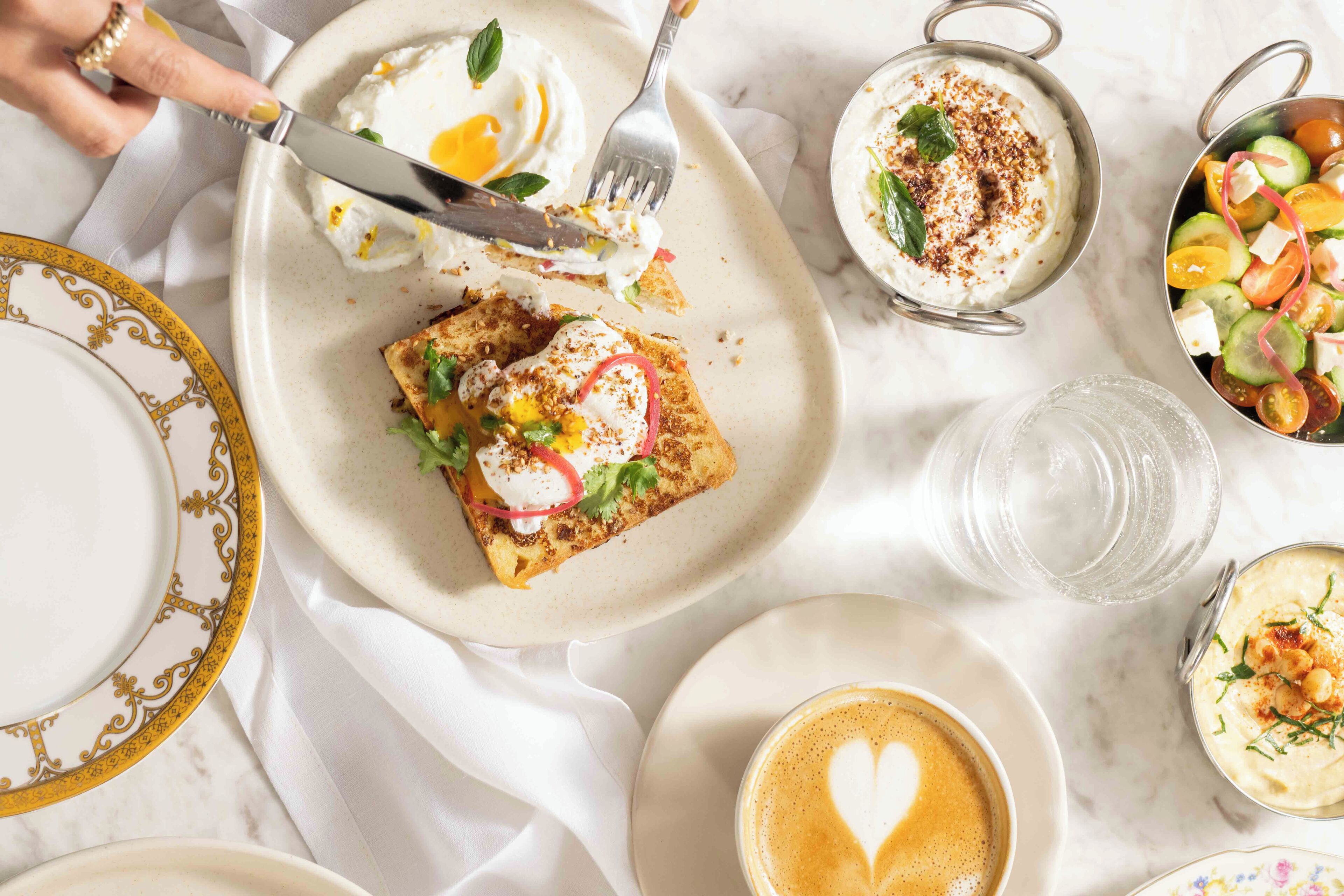 A breakfast setting with a plate of toast topped with egg, a bowl of yogurt, a cup of coffee with a heart-shaped latte art, and a side salad.