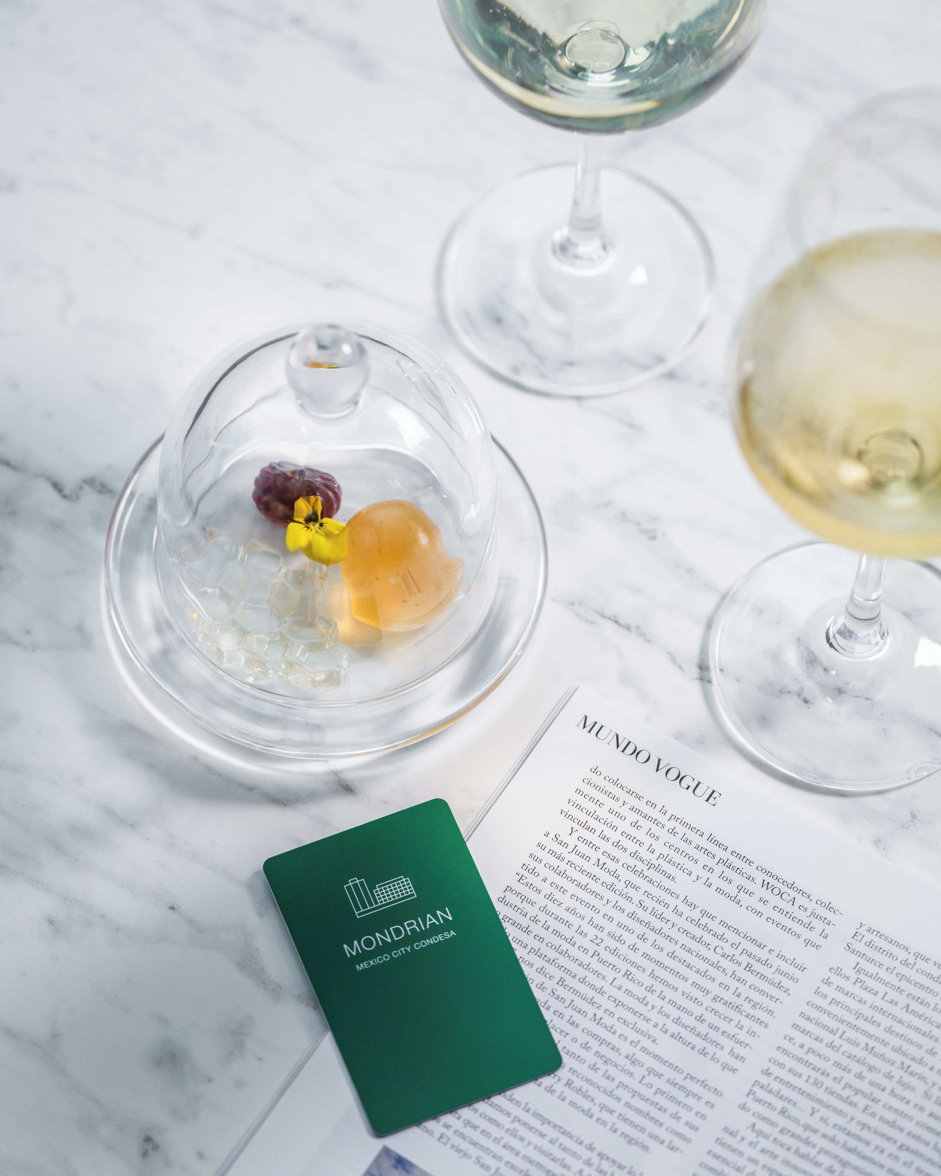A glass dome with a flower and a fruit on a marble surface, a wine glass, and a menu.