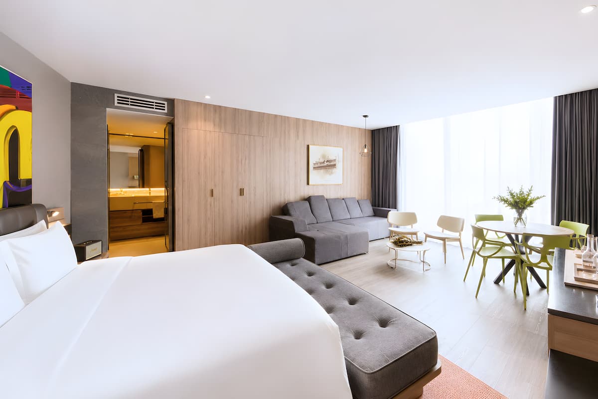 Een hotelkamer met een groot bed, een grijze bank, een eettafel met stoelen en een kleine keukenhoek.