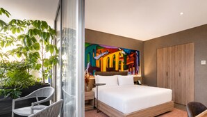 Une chambre d’hôtel moderne dotée d’un grand lit, d’une cloison en verre, d’un coin repas et d’une peinture abstraite aux couleurs vives accrochée au mur.