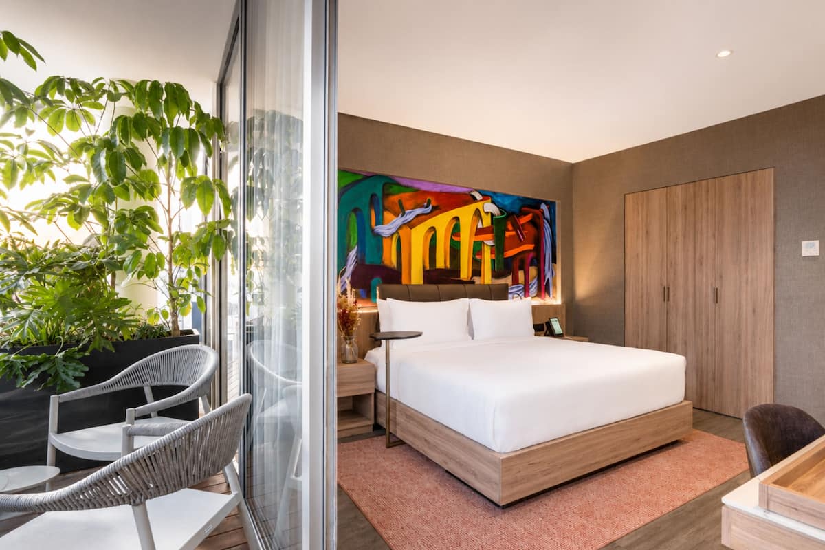 Een moderne hotelkamer met een groot bed, een glazen scheiding, een zithoek en een levendig abstract schilderij aan de muur.