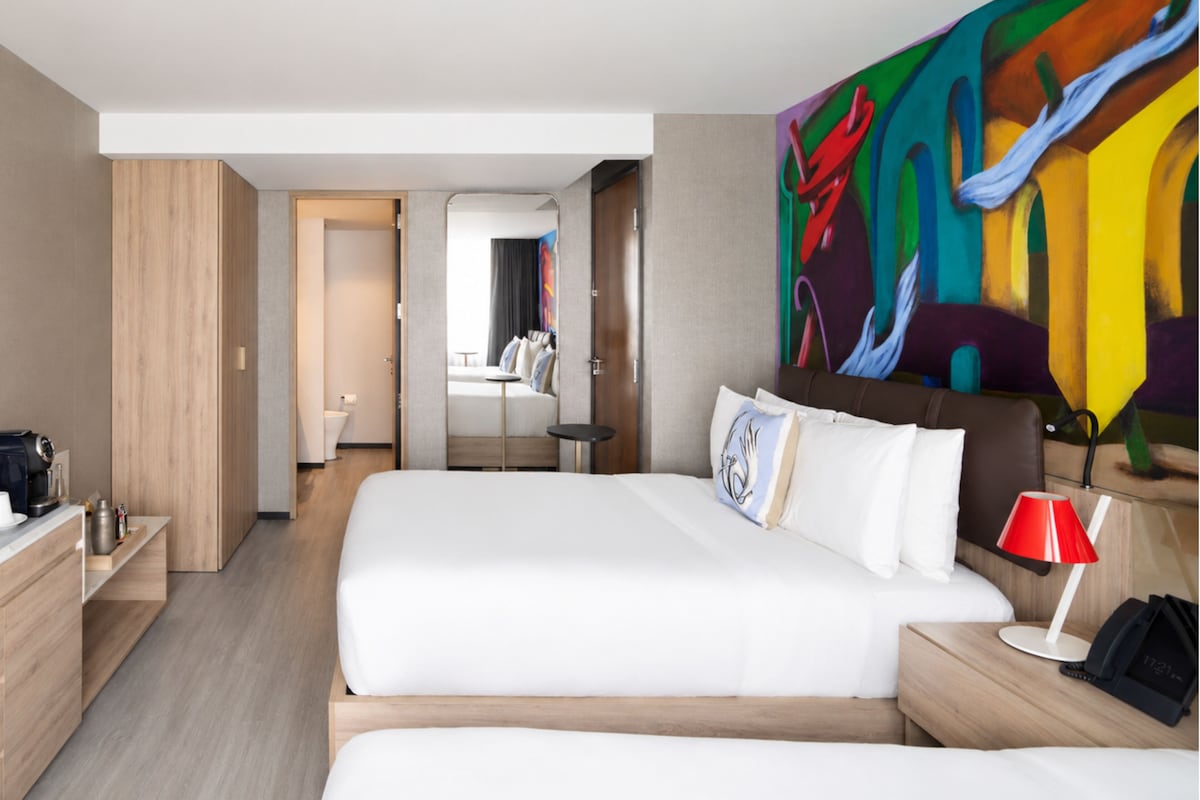 Una habitación de hotel con una cama grande, un escritorio con cafetera, una lámpara roja y un cuadro abstracto colorido en la pared.