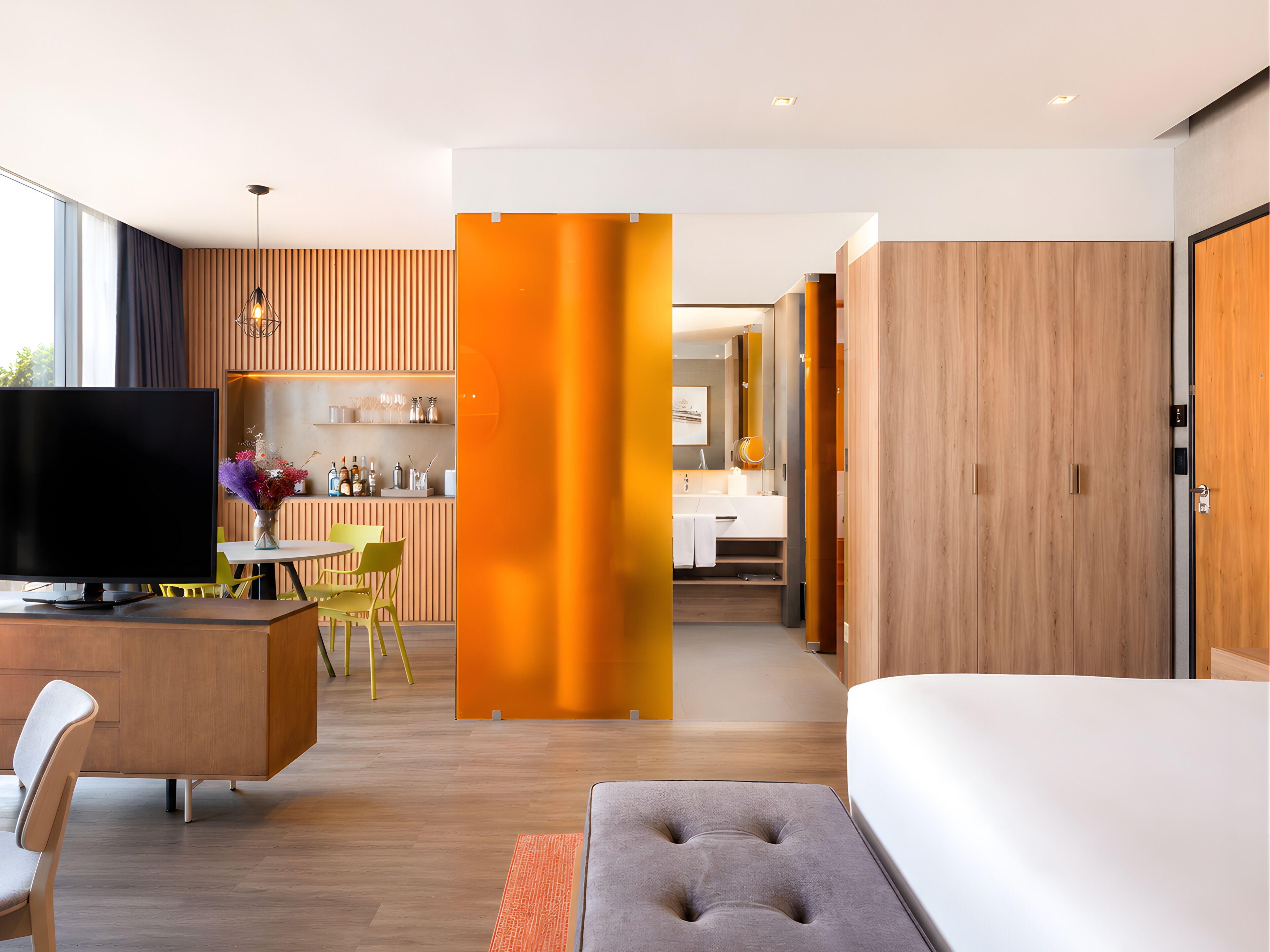 A modern hotel room with a TV, a dining area, a bathroom, and a large bed.