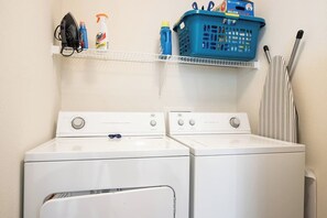 A laundry area with two white washers and dryers, a blue laundry basket, and cleaning supplies on a shelf.