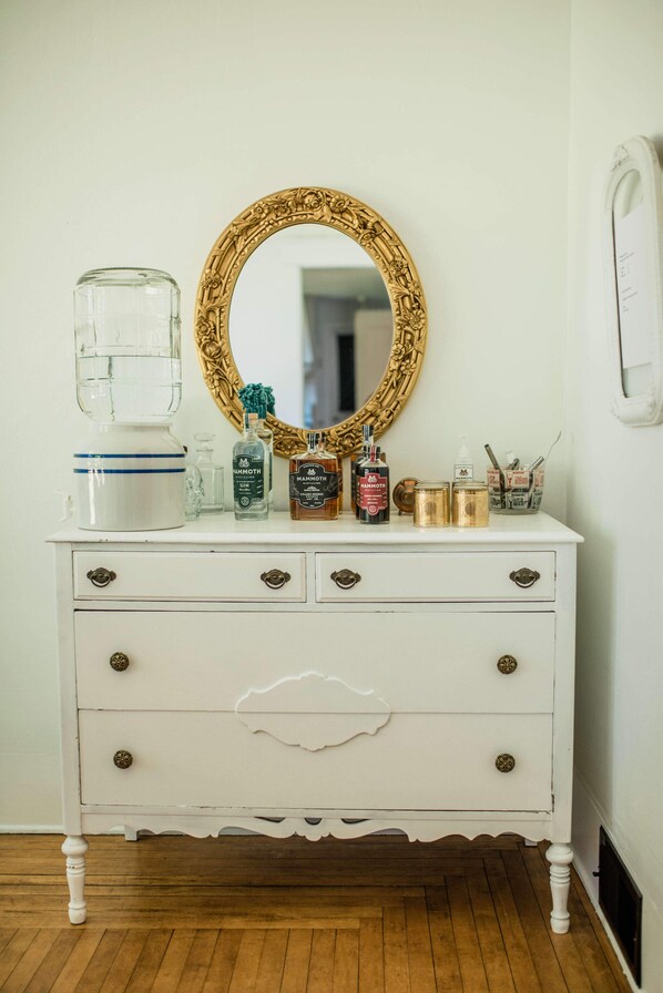 Une commode blanche avec un miroir à cadre doré, des tiroirs, et divers flacons et récipients disposés sur le dessus.