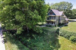Une maison à deux étages avec un porche, entourée d’une végétation luxuriante et d’arbres.