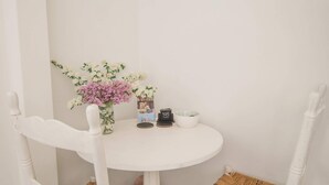 Une petite table blanche d’angle, sur laquelle est posé un vase de fleurs, deux chaises blanches à assise tressée et un tableau encadré accroché au mur.