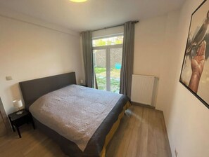 A bedroom with a bed, a nightstand, a window with curtains, and a radiator. - Appartement 3 Chambres Avec Jardin Privatif (Charleroi)