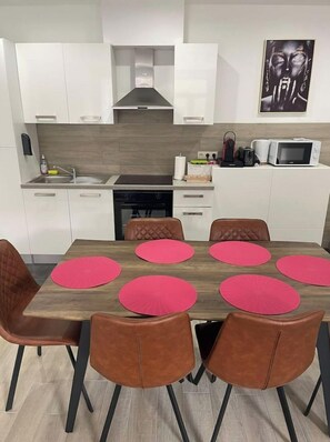 A modern kitchen with white cabinets, a microwave, and a sink. There is a dining table with six chairs and pink placemats. - Appartement 3 Chambres Avec Jardin Privatif (Charleroi)