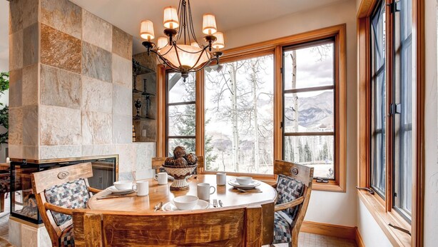 A cozy dining area with a wooden table, chairs, and a chandelier. Large windows offer a view of trees and mountains. - Luxury Lodge with Breathtaking Views (Mountain Village)