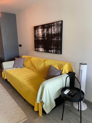 Ein modernes Wohnzimmer mit einem gelben Sofa, einer grau-weißen Decke, einem schwarz-weißen abstrakten Gemälde, einem Heizlüfter und einem kleinen schwarzen Beistelltisch mit Fernbedienung.