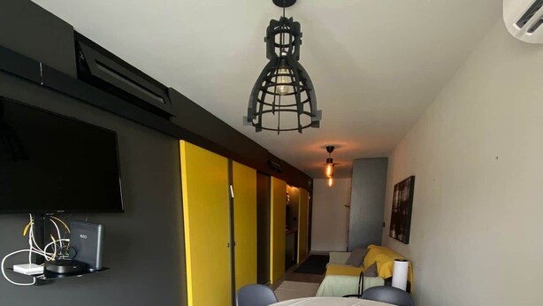 Een eetruimte met een ronde tafel, stoelen, een plafondlamp en een aan de muur bevestigde televisie.