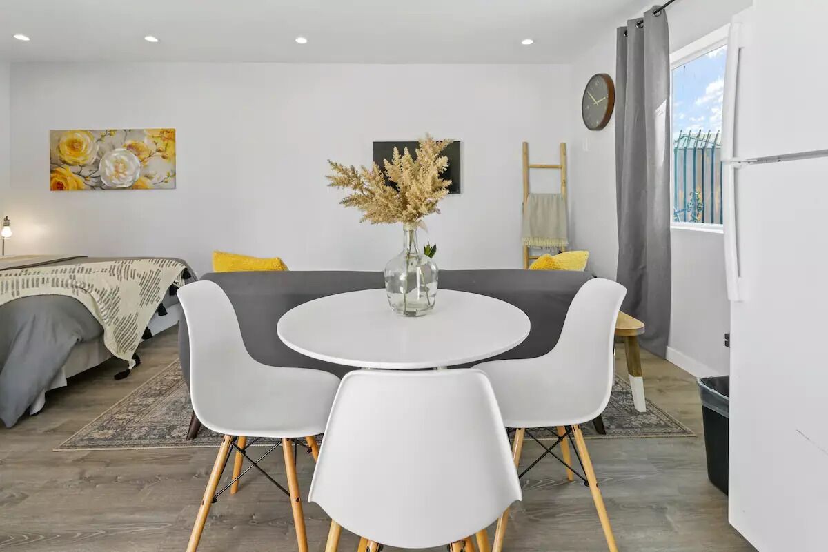 A modern, compact living space with a round dining table, white chairs, a sofa with yellow cushions, a large window with a view, and a decorative vase with dried flowers on the table.
