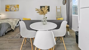 A modern, compact living space with a round dining table, white chairs, a sofa with yellow cushions, a large window with a view, and a decorative vase with dried flowers on the table. - Adorable studio close to DTLA (Alhambra)