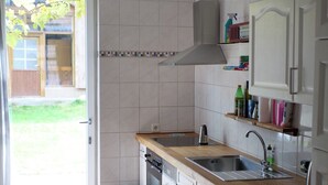 Une cuisine dotée d’un plan de travail en bois, d’un évier en acier inoxydable et de meubles blancs. On y trouve une poubelle et une fenêtre donnant sur une cour verdoyante.
