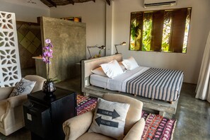 A bedroom with a bed, bedside tables, a chair, a decorative plant, and a window with a view of greenery. - Casa Na Lagoa (Paracuru)