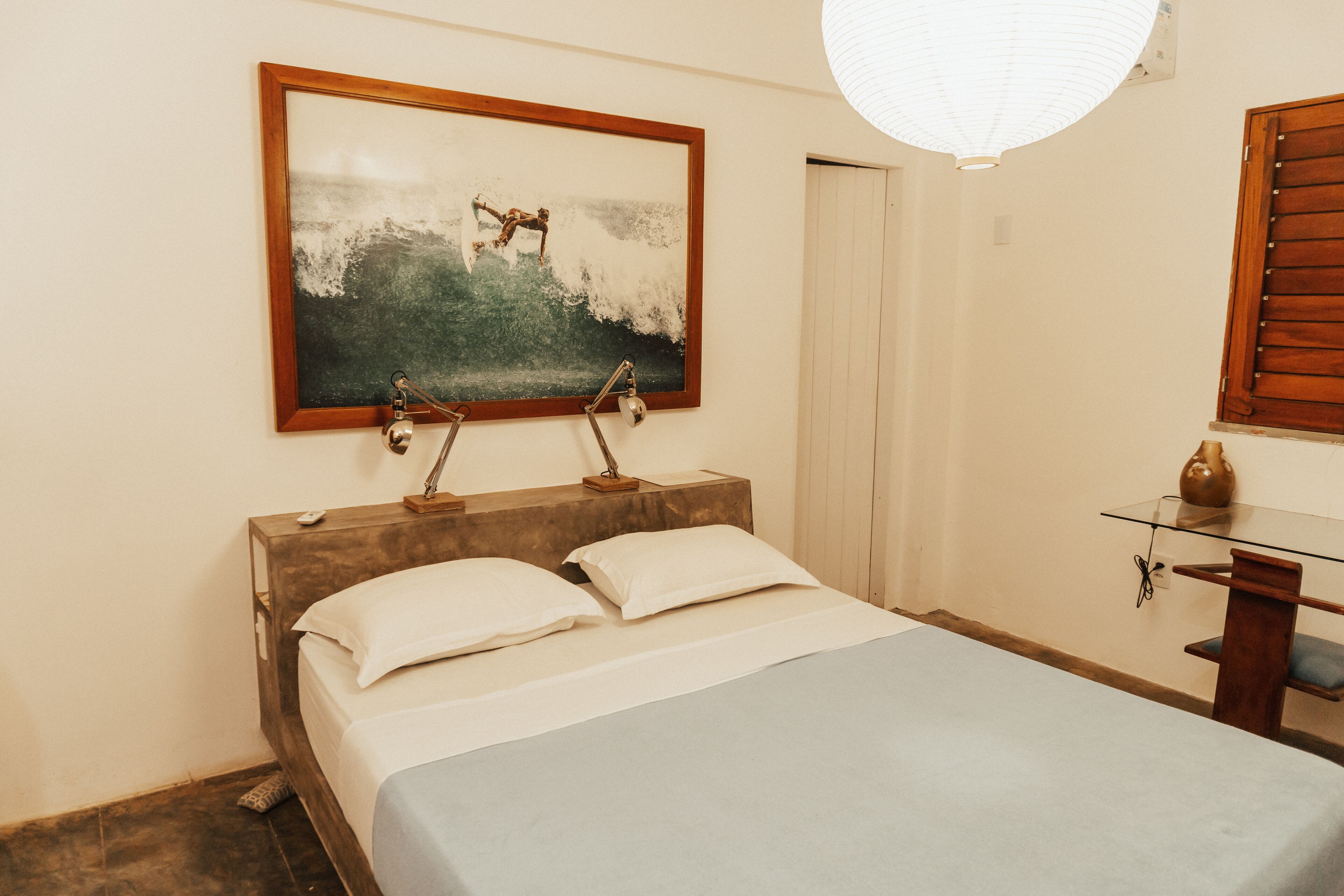 A bedroom with a bed, bedside lamps, a painting, a desk with a vase, and a window with blinds.