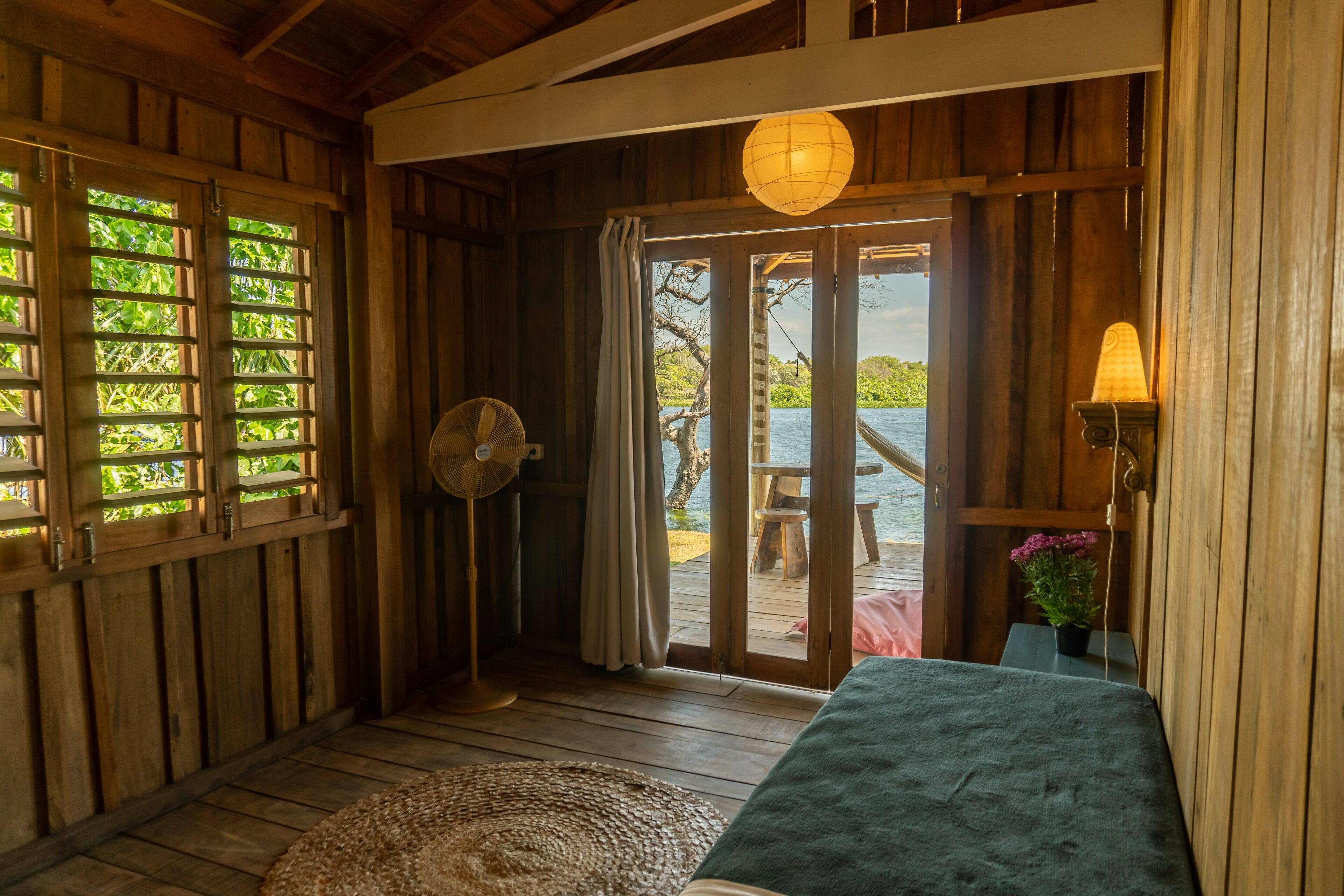 Habitación de madera con cama, ventilador y vista a una masa de agua a través de un vidrio.