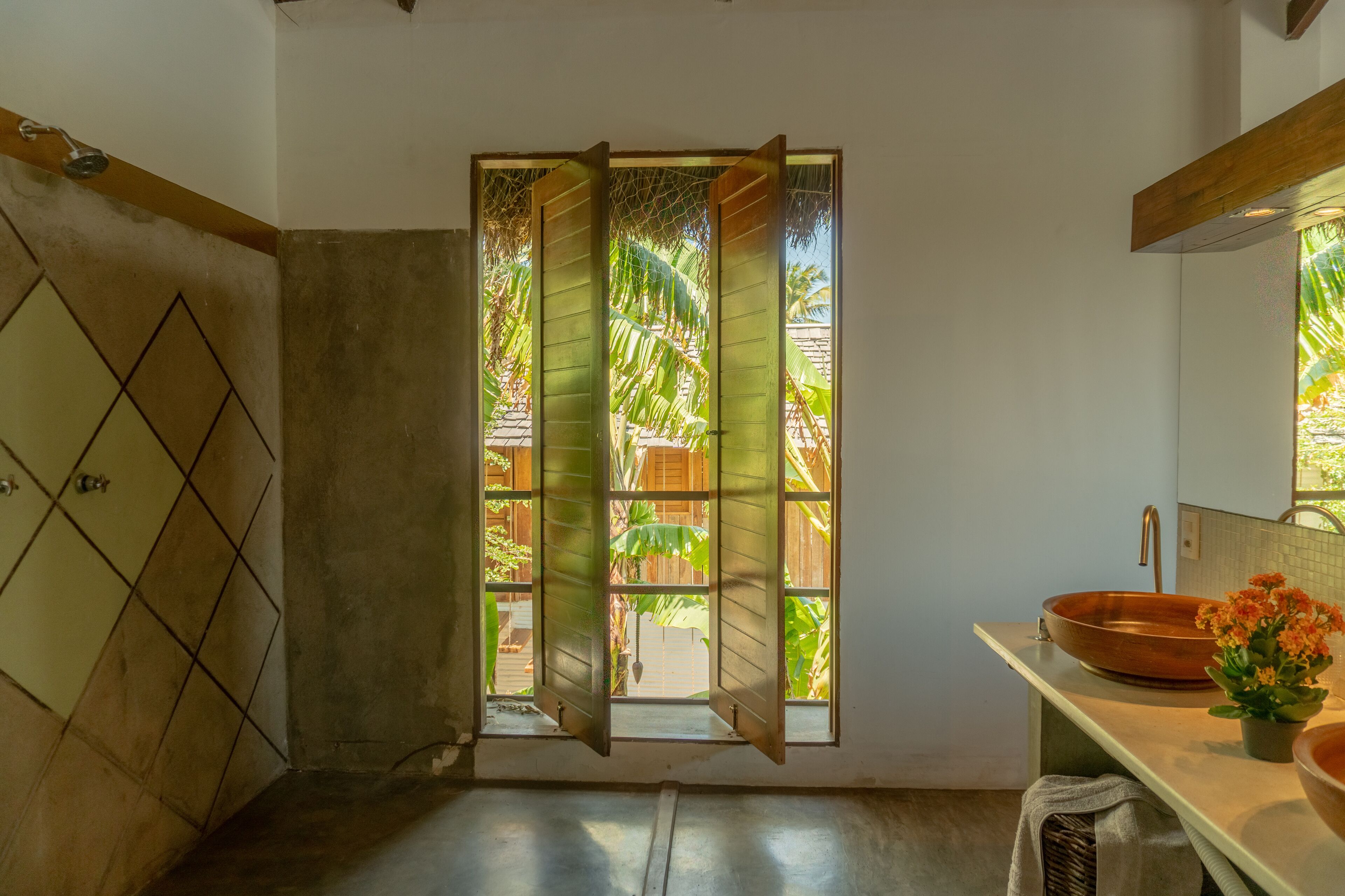 Un baño con un ventanal grande, una puerta de madera, un lavabo con cuenca de cobre y una planta en maceta.