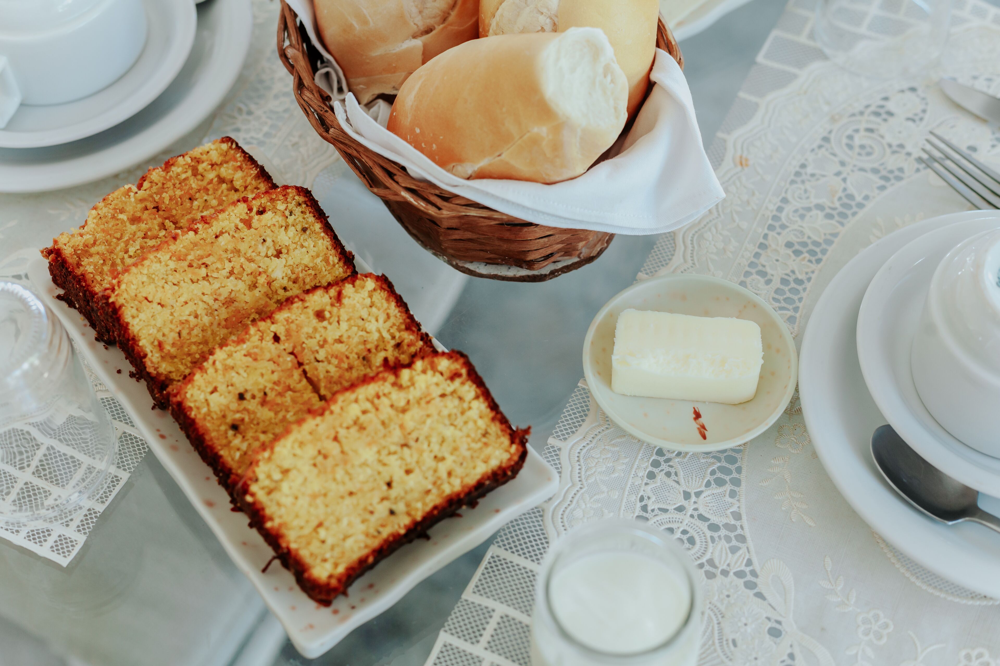 Una mesa adornada con un mantel de encaje blanco, un plato de pastel, una cesta de bollos y un tazón pequeño de mantequilla.