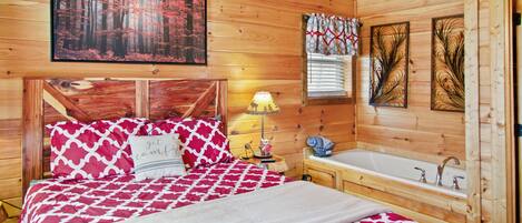 A wooden cabin bedroom with a bed, a nightstand, a lamp, a window with blinds, and a wooden bathtub.
