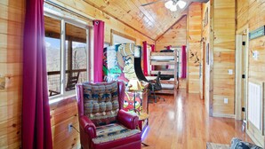 Interior de uma cabana de madeira com uma cadeira vermelha, um ventilador de teto e uma janela grande com cortinas.