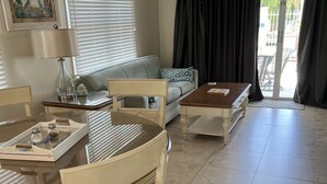 Un salon comprenant une table à manger, des chaises, un canapé et une table basse. Il y a une fenêtre avec des stores, une porte avec une pancarte et une vue sur l’extérieur.