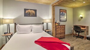 Een hotelkamer met een houten plafond, een bed met witte lakens, een rode plaid, een bureau met stoel, en twee wandlampen.
