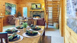 Interno di una baita in legno con zona pranzo apparecchiata per quattro persone, camino e una scala di legno che conduce a una cascata.