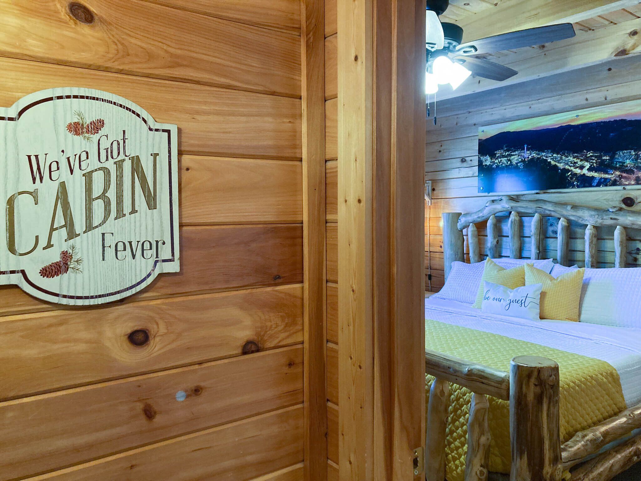 Een houten cabin interieur met een bed, een bordje met de tekst 'We' Got CABIN Fever' en uitzicht op een stadsgezicht door het raam.