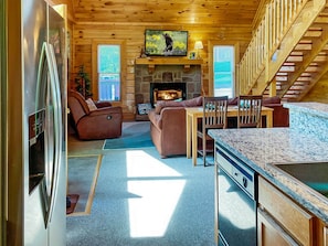 L’intérieur chaleureux d’un chalet avec une cheminée en pierre, des meubles en bois, un coin repas et une cuisine équipée d’un réfrigérateur.