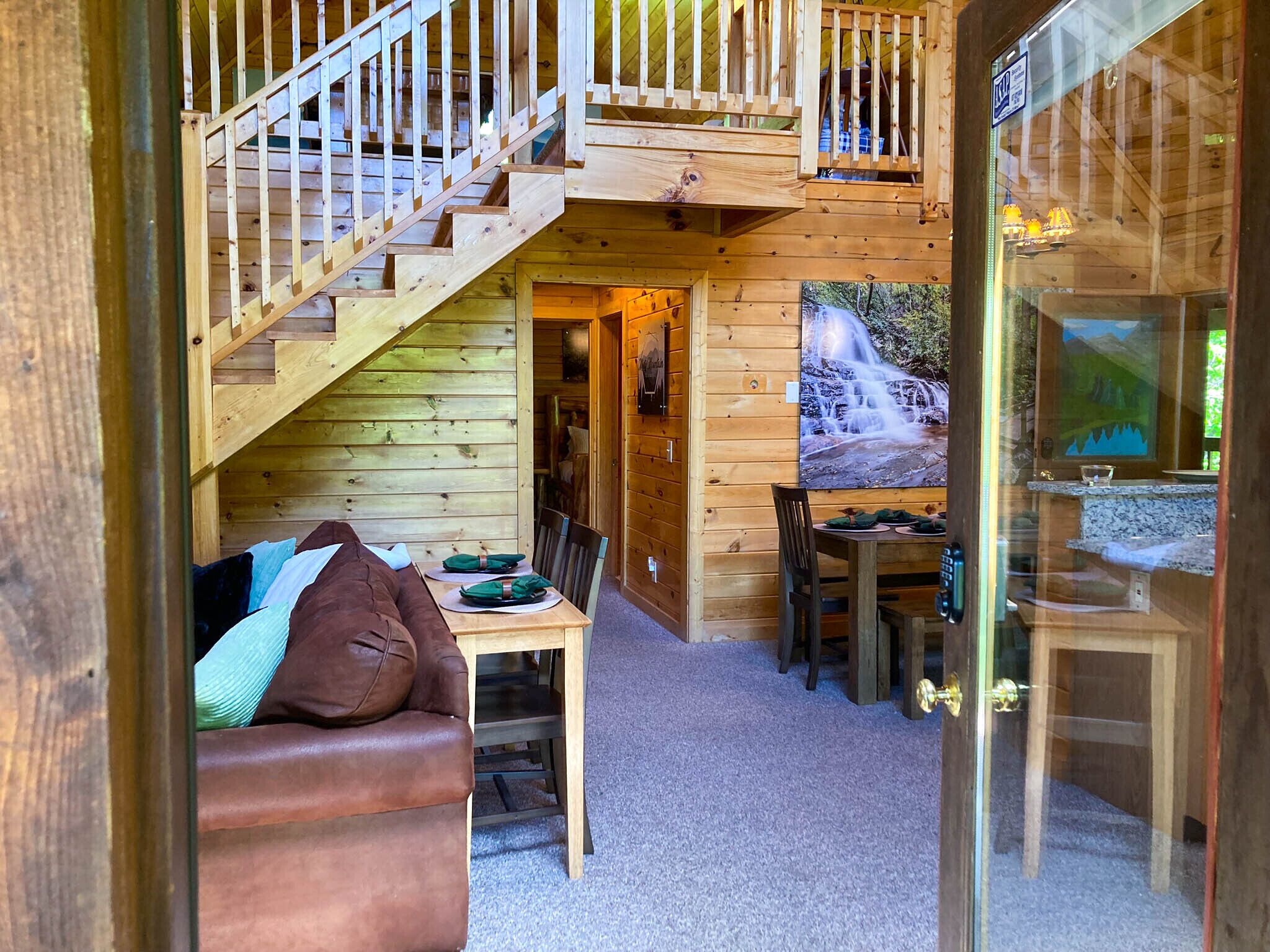 Interior d'una cabana de fusta amb una escala que porta a un espai al costat, una zona de menjador amb taula i cadires, i una finestra gran amb vista a una cascada.