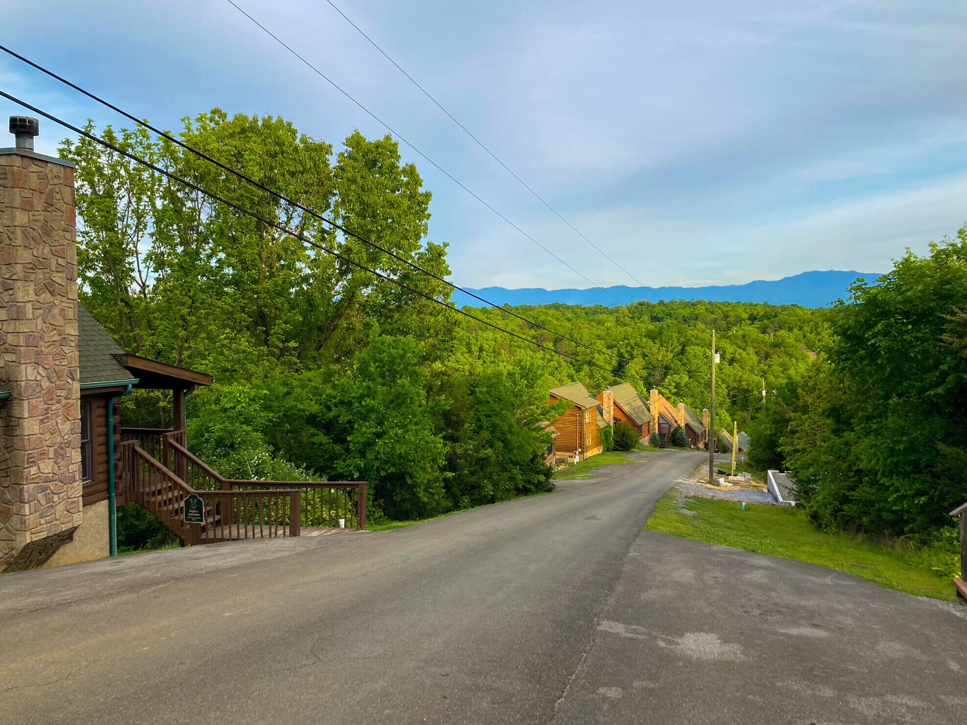 A rural road leading to a stone building with a porch, surrounded by lush greenery and mountains in the background.