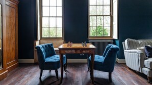 A room with a wooden table, two blue armchairs, a white-striped sofa, and two windows with a view of greenery. - Beautiful 7 bedroom house hottub wifi (Shepton Mallet)