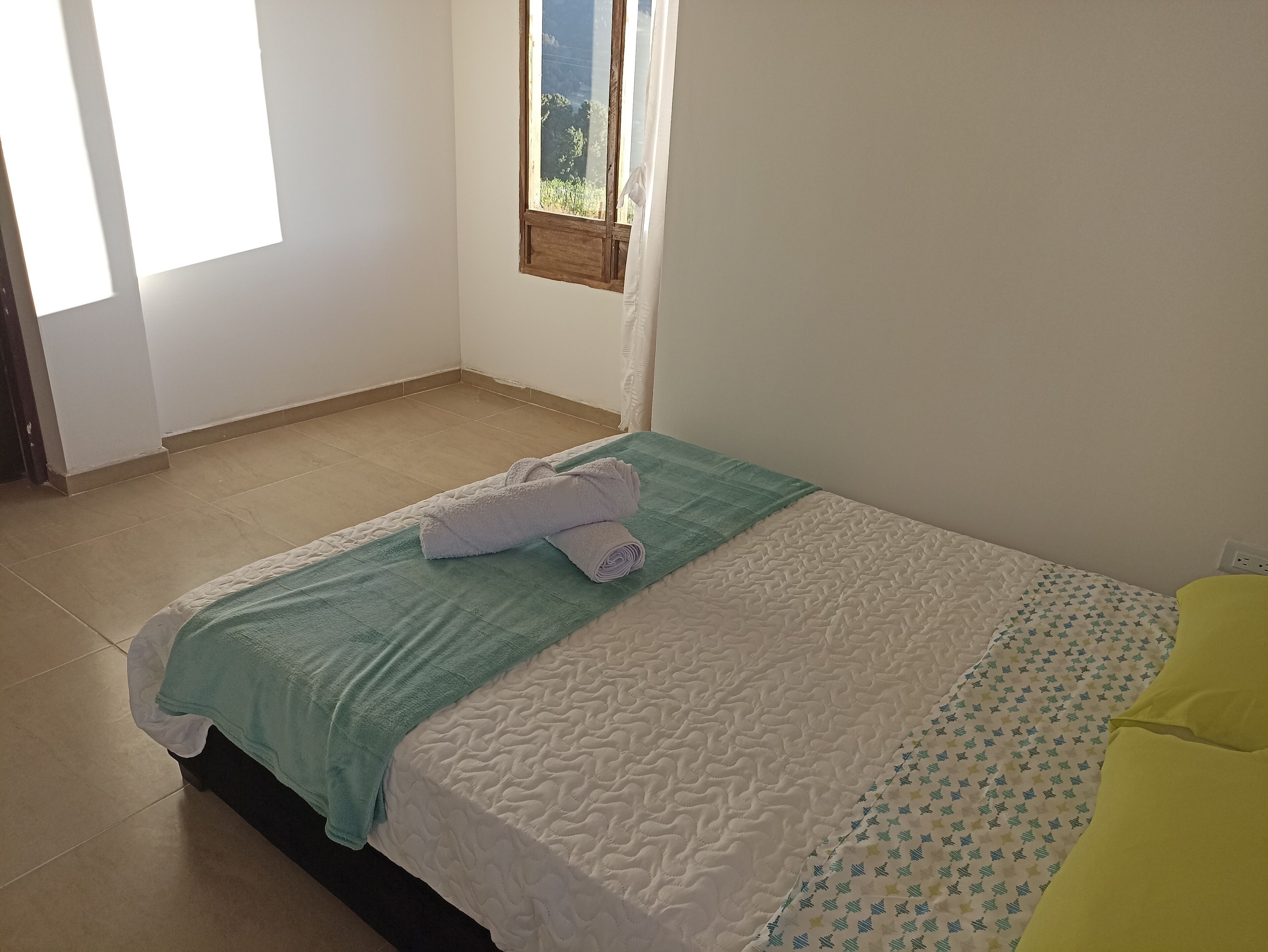 A neatly made bed with a green and white quilt, a rolled white towel, and a yellow pillow.