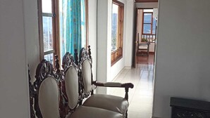 A room with a wooden ceiling and white walls. There are two ornate wooden chairs with cushioned seats and backs. A small wooden bench is against the wall. The room has large windows with curtains, and a view of a balcony is visible through the windows. - Hotel Tota Eliseo Playa Blanca (Tota)