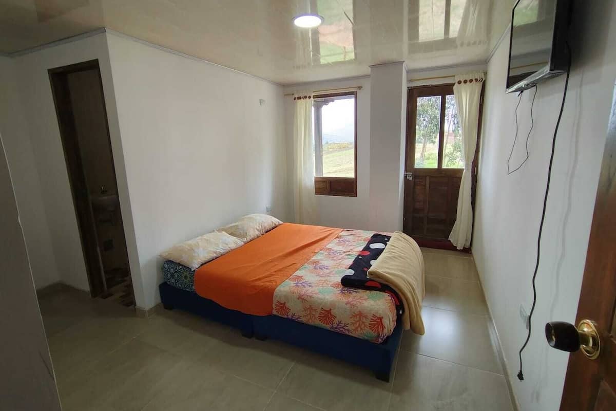 Un dormitorio con una cama, un televisor montado en la pared y una ventana con cortinas.