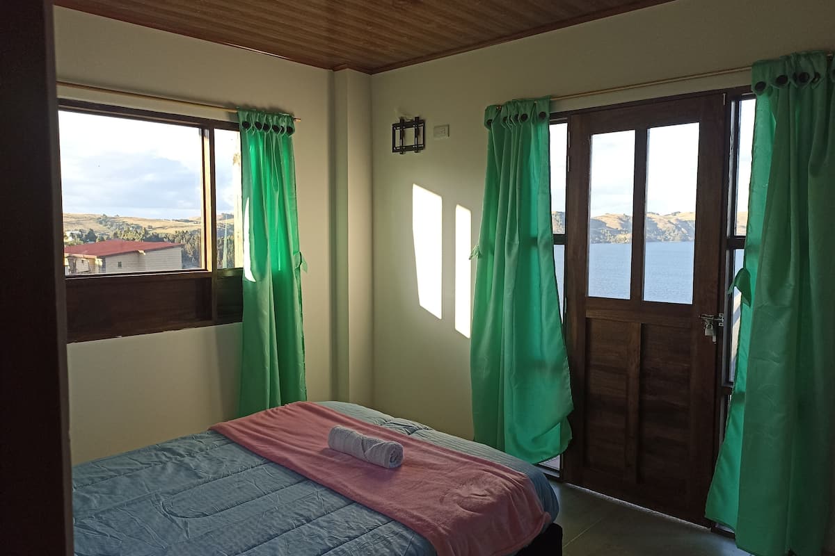 Una habitación con cama, ventana con cortinas, puerta y vista a un cuerpo de agua y colinas.