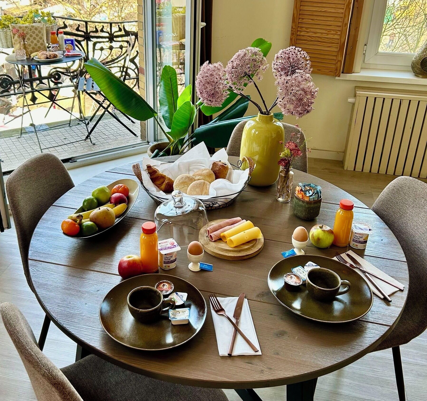A dining table set with breakfast items, including fruits, bread, and beverages, in a room with a view of the outdoors.
