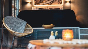 Una habitación acogedora con una silla de mimbre, un sofá negro, un cuenco de fruta y una vela decorativa.