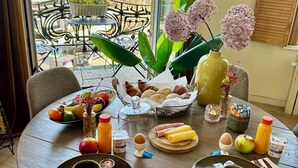 Una mesa de comedor con alimentos para el desayuno, un florero con flores y vistas a edificios a través de una ventana.