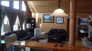 Een gezellig chaletinterieur met houten wanden, een houten plafond, een eethoek met tafel en stoelen, een open haard en een groot raam.