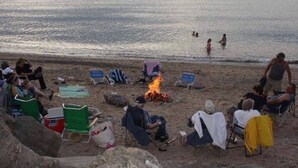 Een strand met mensen die zitten en staan, een vuurkuil en uitzicht op de zee.