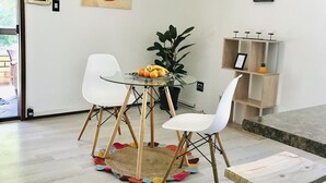 A modern living room with a glass table, white chairs, a wooden stool, a potted plant, and a wooden shelf with decorative items.