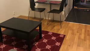 Ein schlichter, wenig möblierter Raum mit Holzboden, einem schwarzen Tisch, zwei schwarzen Stühlen und einem roten Teppich mit Muster.