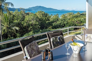 Ein Tisch mit einer Weinflasche, Gläsern und Fernglas auf einem Balkon mit Blick auf eine üppige Landschaft, das Meer und ferne Berge.
